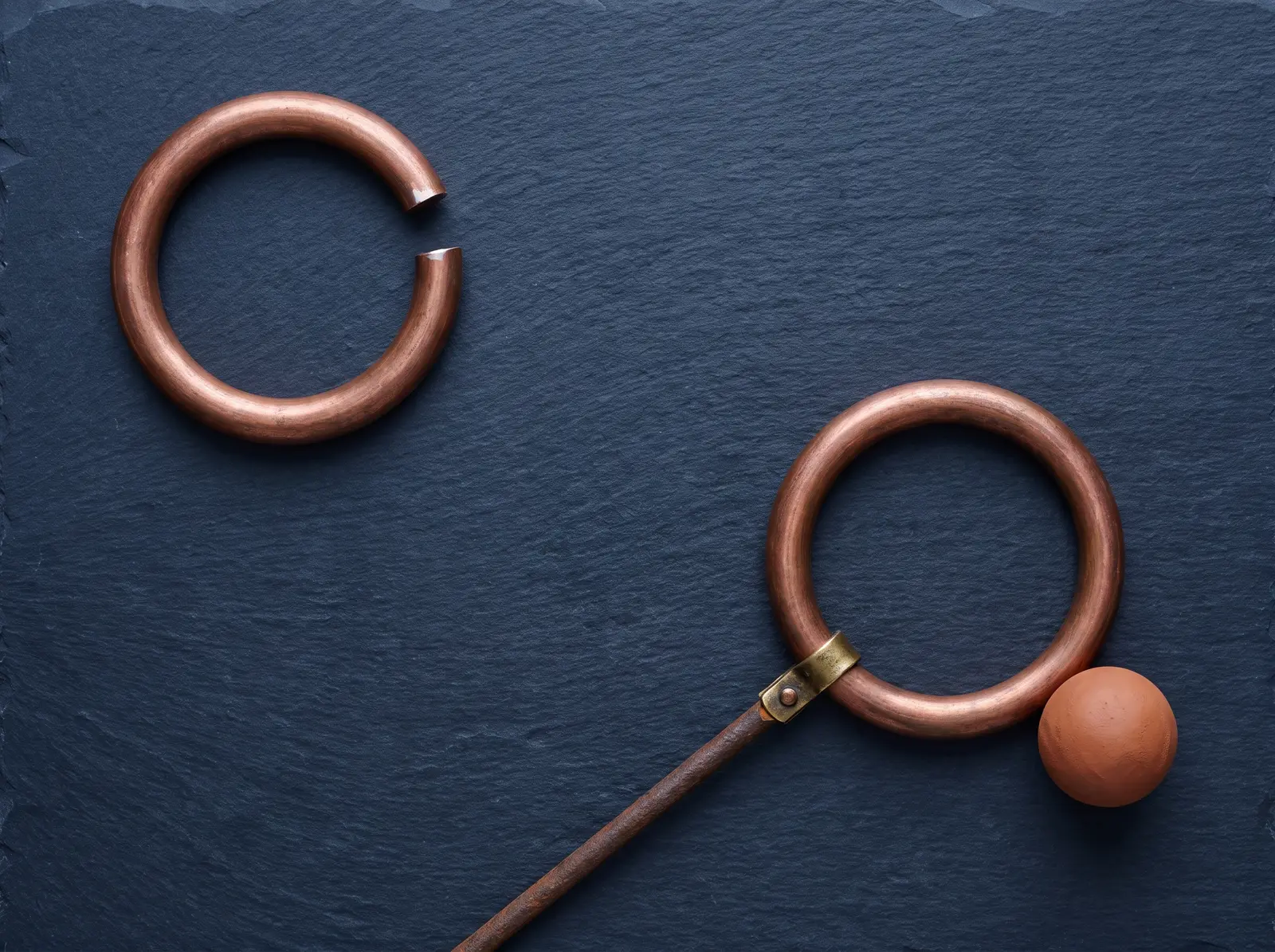 Still life on dark slate: one copper ring standing open with a gap cut through it, and a second copper ring closed, held to a rusted rod by a brass clasp with a small clay sphere resting at its rim